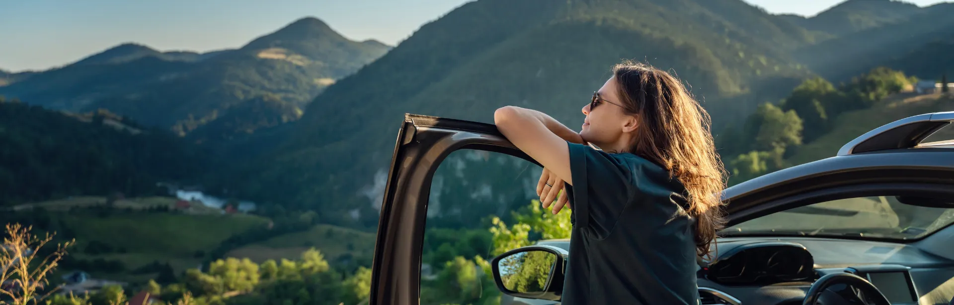 A woman rests her arms on a car door in a mountainous area.