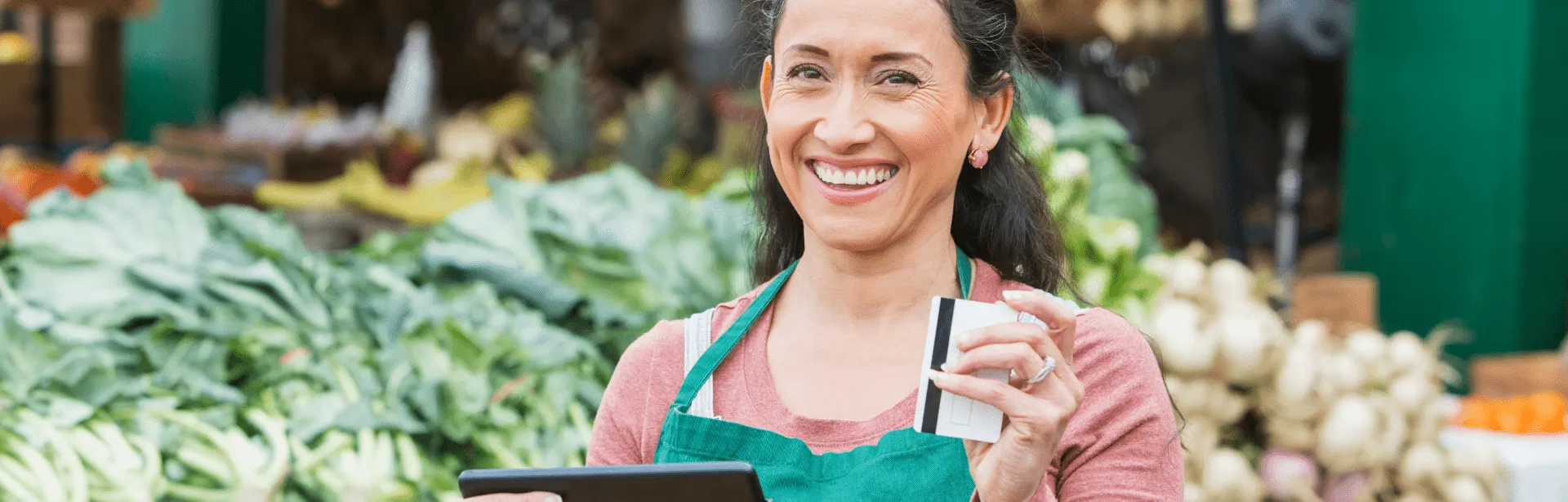 A woman in a produce section of a grocery store wearing an apron and holding a debit card.