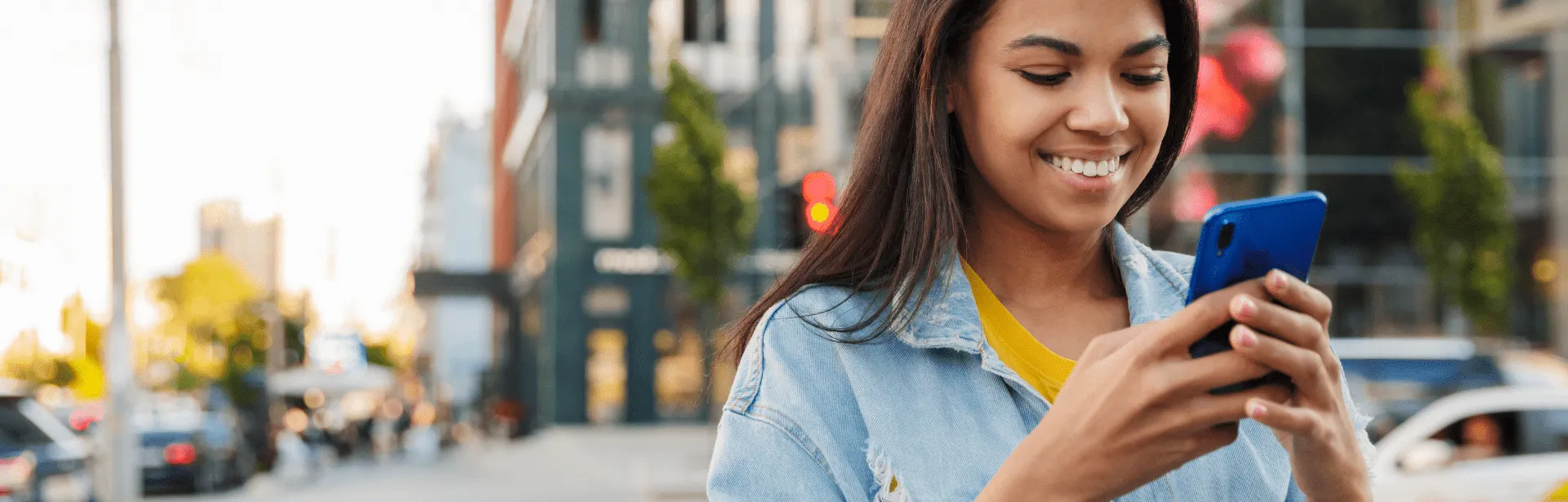 A woman walking down the street in an urban area smiling and looking at phone screen.